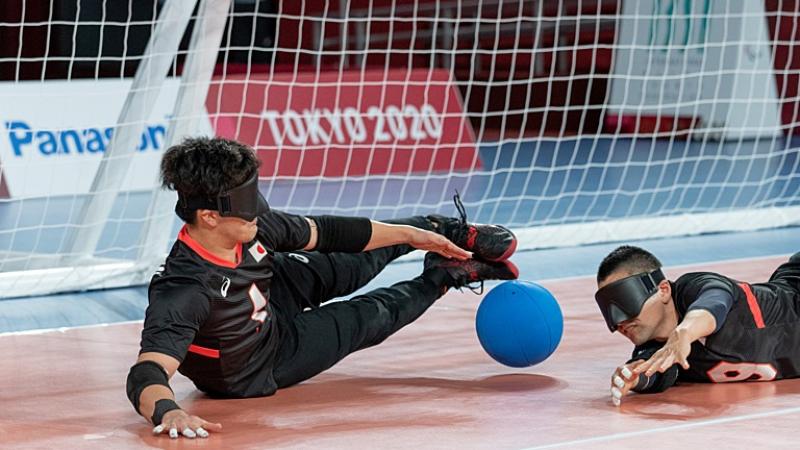 Two male goalball athletes are blocking the ball in front of the goal. They have their hands on the court.