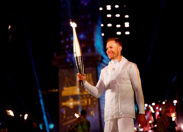 Markus Rehm, a male long jump athlete, wears a white uniform and holds the Paris 2024 torch during the Paris 2024 Paralympic Opening Ceremony