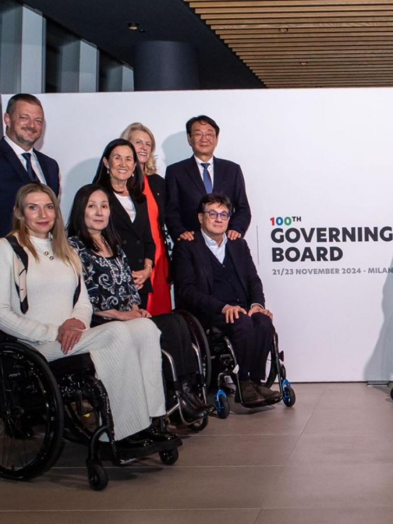 Fourteen people wearing formal clothing are posing in front of a banner that says "100th Governing Board"