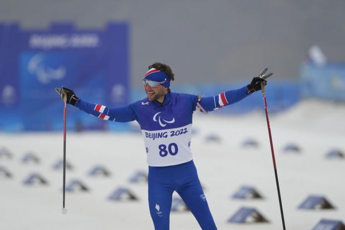A male athlete wearing a blue skiing gear and skiing goggles sticks celebrates by raising both arms on the snow