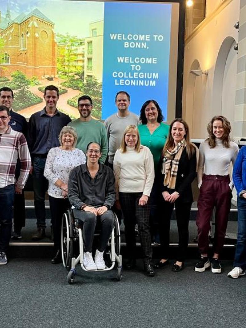 19 people pose for a photograph in front of a screen that says "Welcome to Bonn"