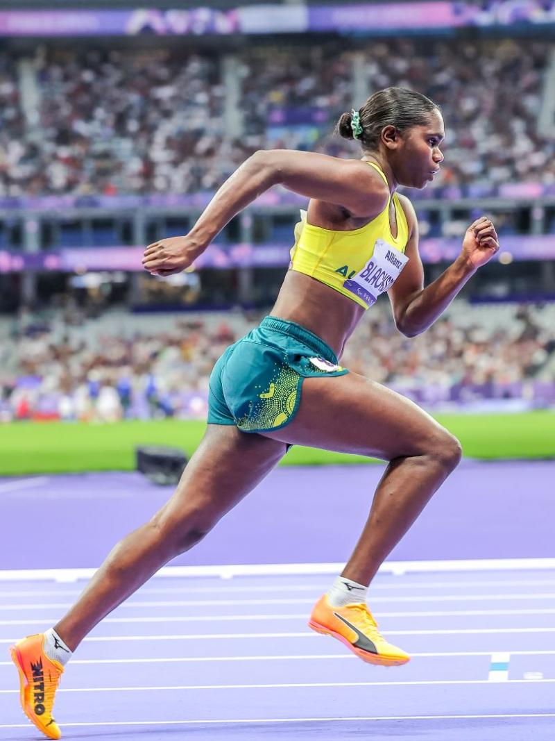 An Australian female athlete with a yellow vest and green shorts runs at the Stade de France athletics stadium