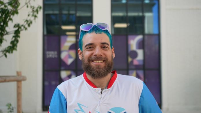 A male athlete with blue hair and heart-shaped sunglasses smile at the camera