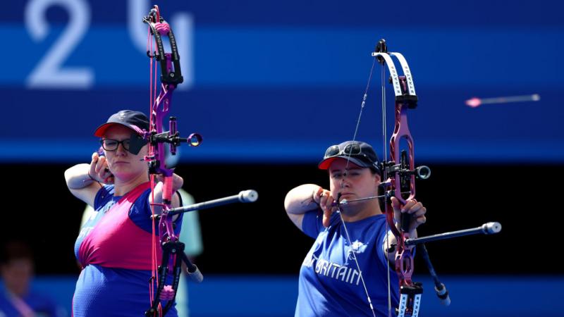 Jodie on the left in pink and blue with an obvious baby bump and Phoebe Paterson Pine on the right in blue Great Britain t-shirt both raise their bows to eye level to line up a shot