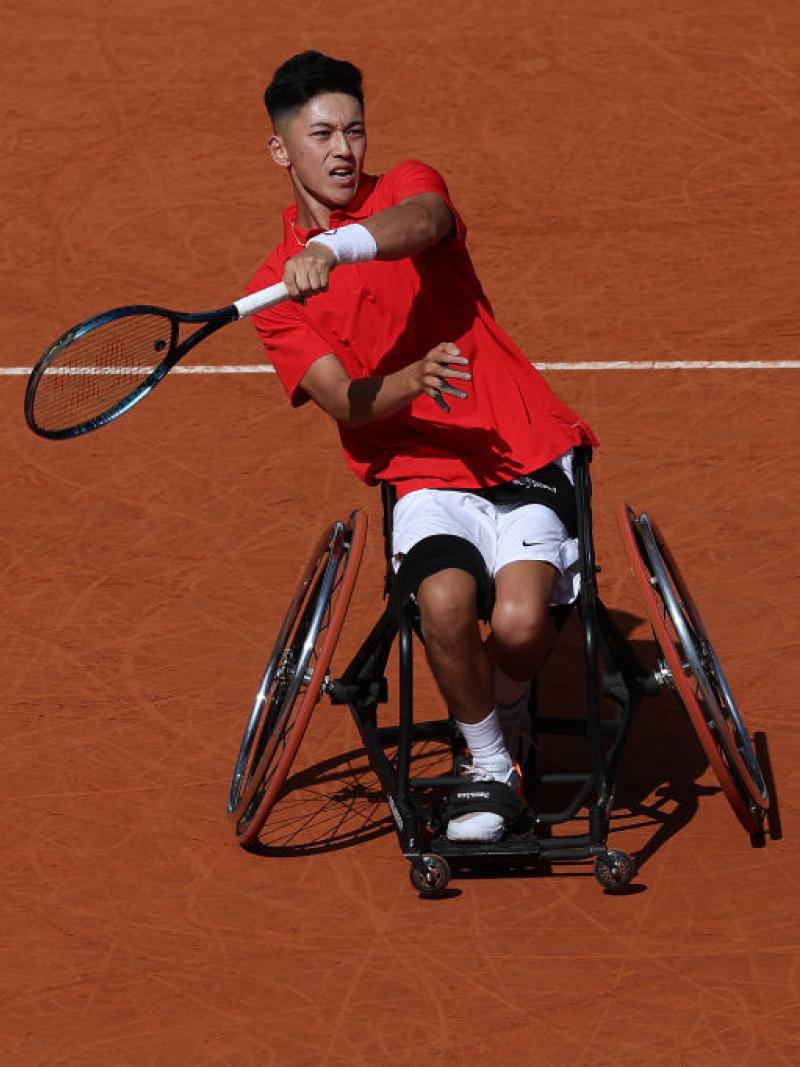 Against an orange earth background a man in a red tee and a wheelchair reaches a racket out as a yellow tennis ball flies