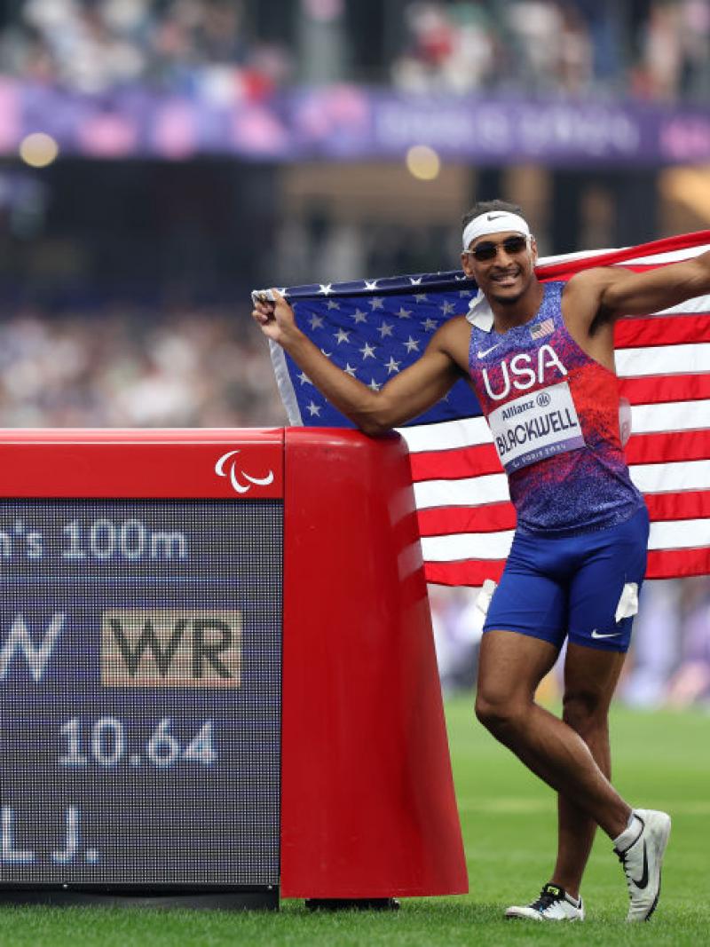 A man in a Team USA uniform holds up an American flag and stands next to an LED screen that shows his new world record time