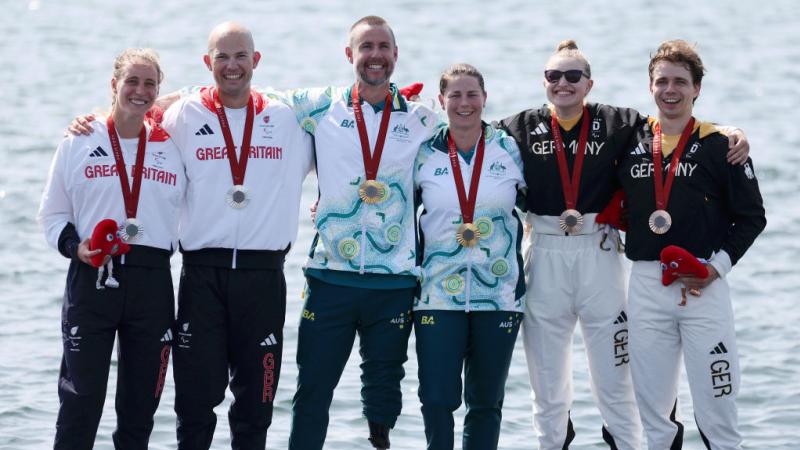 Six athletes stand on a podium next to water, two with silver medals, two with gold medals, and two with bronze medals