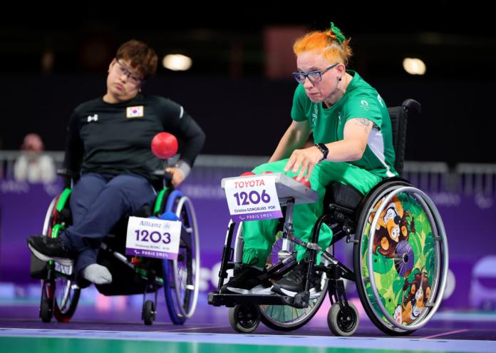 Two women in wheelchairs play boccia. One woman in green throws a red ball.