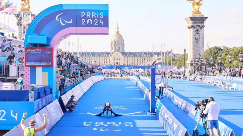Bike crosses the para triathlon finish line with rider leaning forward showing a white helmet, surrounded by Paris 2024 branding