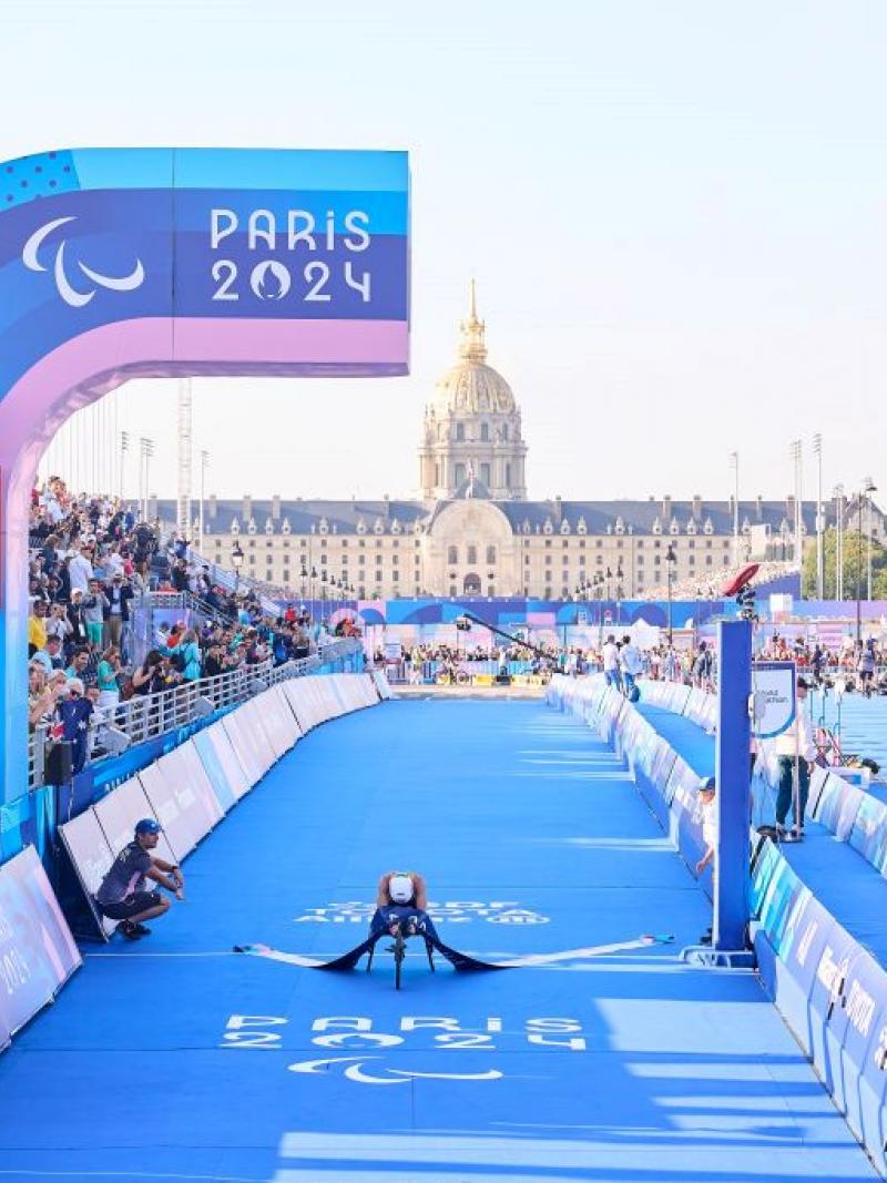 Bike crosses the para triathlon finish line with rider leaning forward showing a white helmet, surrounded by Paris 2024 branding