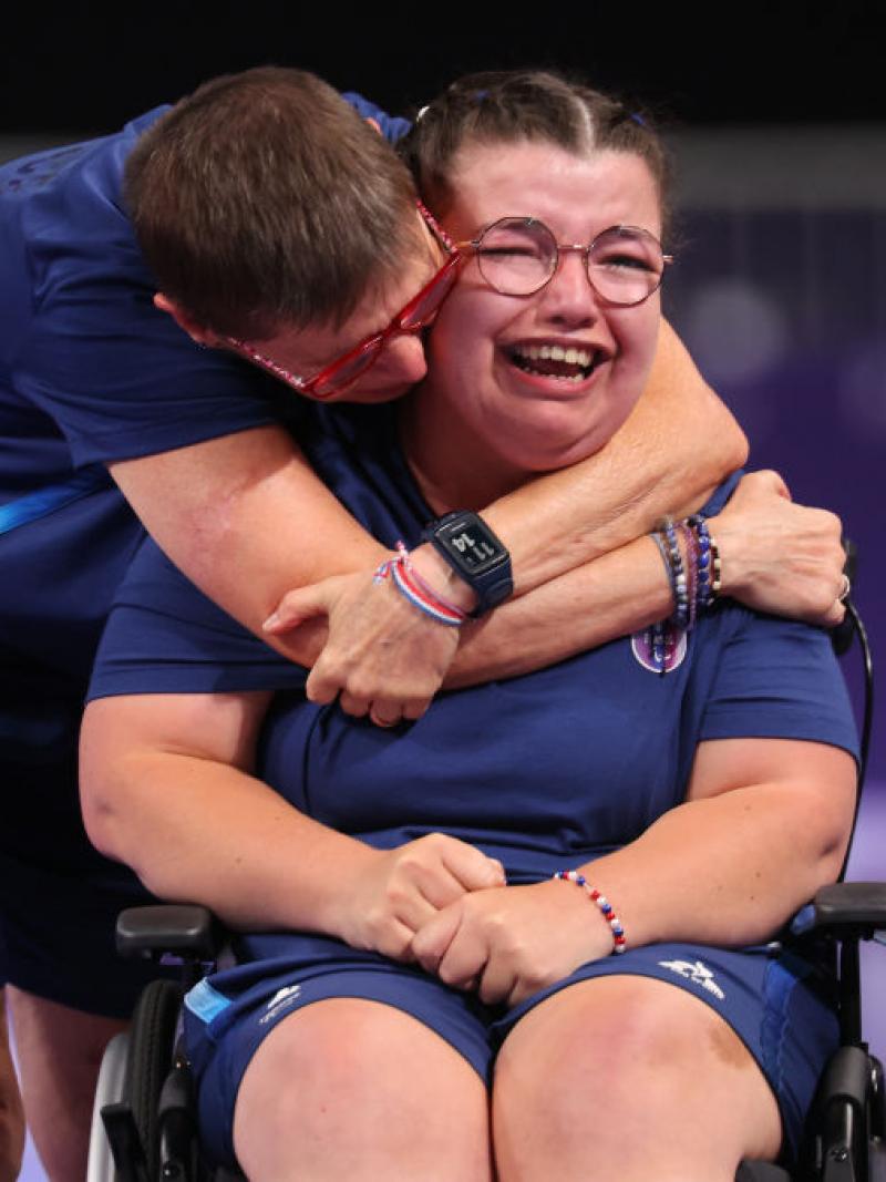 A woman in blue tee and shorts seated in wheelchair is hugged by another woman while crying