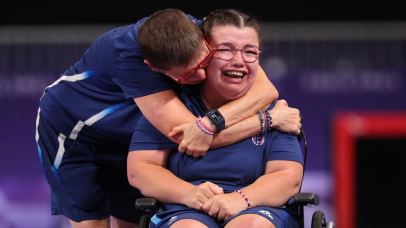 A woman in blue tee and shorts seated in wheelchair is hugged by another woman while crying