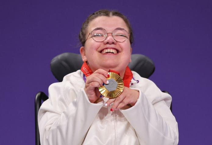 A woman in a white jacket and a wheelchair closes her eyes and smiles while holding up a gold medal