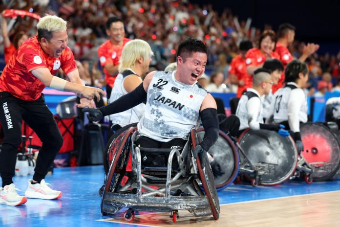 A man in a wheelchair yells happily. He is on a wheelchair rugby court. More people in wheelchairs behind him smile, the crowd in the stands cheers.