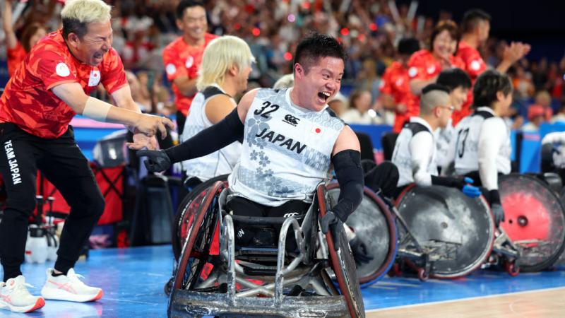 A man in a wheelchair yells happily. He is on a wheelchair rugby court. More people in wheelchairs behind him smile, the crowd in the stands cheers.