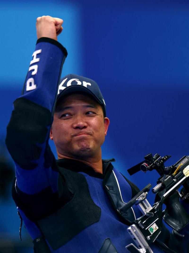 A man in a dark blue jacket and baseball hat lifts one arm in the air in celebration and holds his shooting Para sport rifle in his other hand.