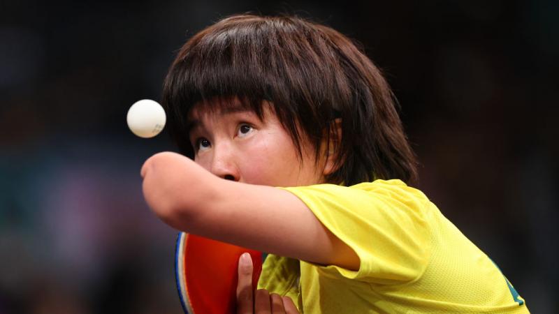 A table tennis player in a yellow shirt keeps her eye on the ball as she serves