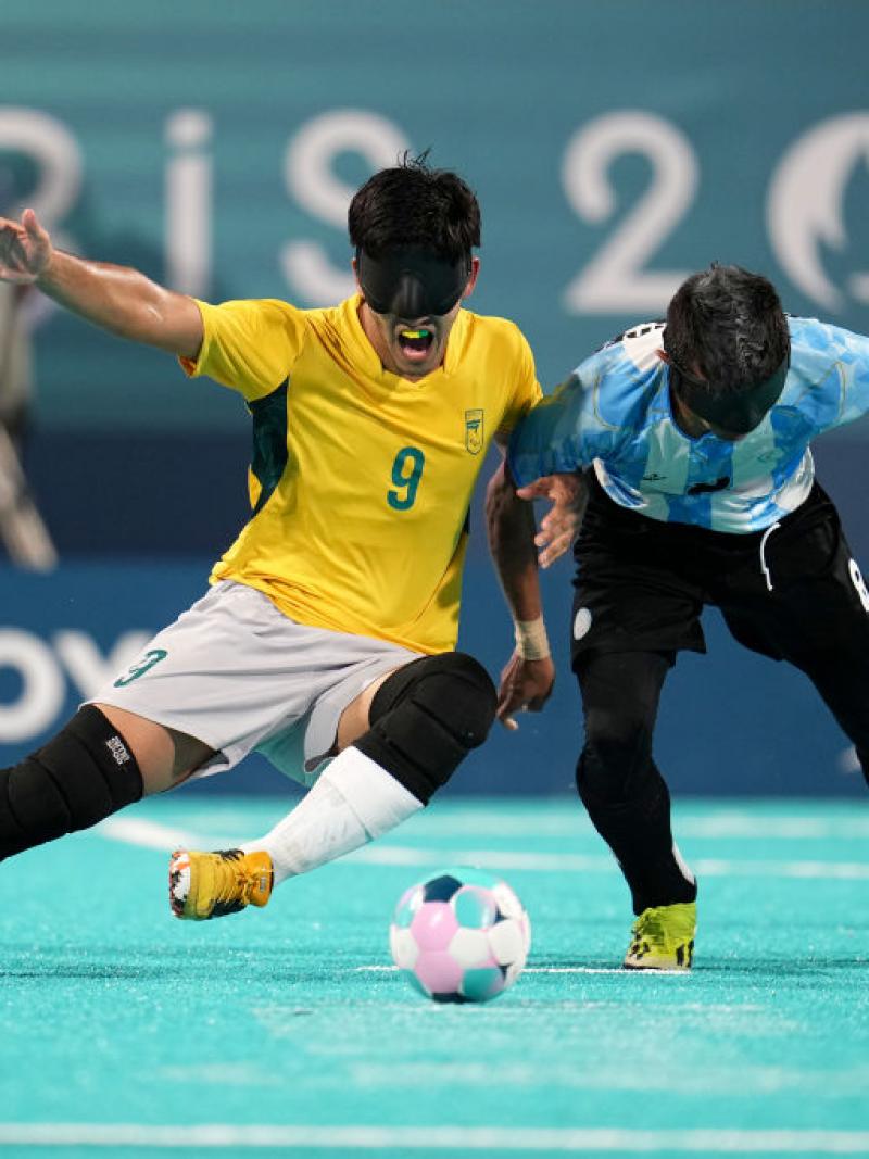 Two blind football players wearing black eyeshades, one in a yellow Brazil jersey and one in a blue and white Argentina jersey, compete for the ball