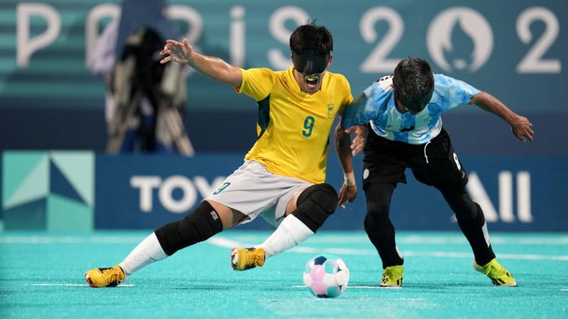 Two blind football players wearing black eyeshades, one in a yellow Brazil jersey and one in a blue and white Argentina jersey, compete for the ball