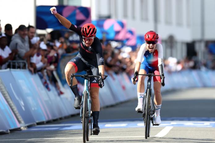 Two women cyclists, one on the left just ahead raises her hand in celebration