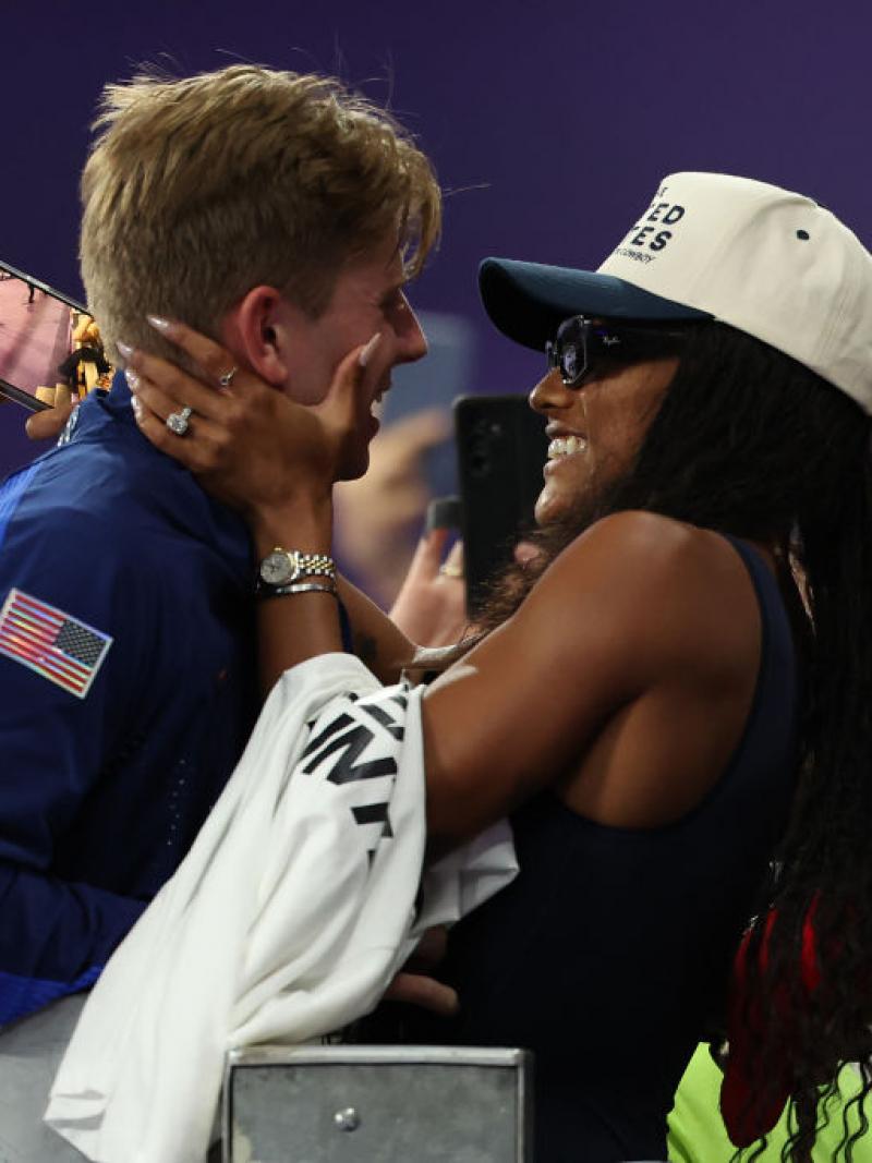 A woman in the crowd in a United States baseball hat puts her arms around a man in a Team USA jacket, both smiling
