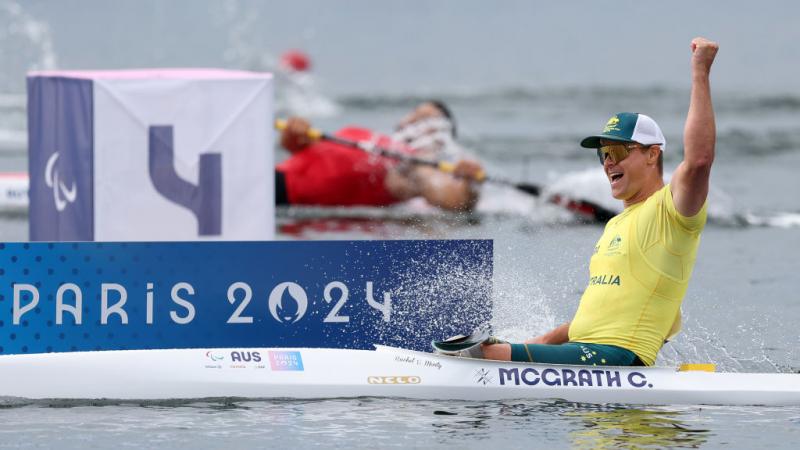 An Australian male rower wearing a yellow shirt, sunglasses and a green hat, celebrates a rowing win by punching the air in his boat