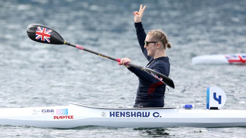 A woman in a canoe holds her oats with one hand and lifts the other hand in the air, making a peace sign