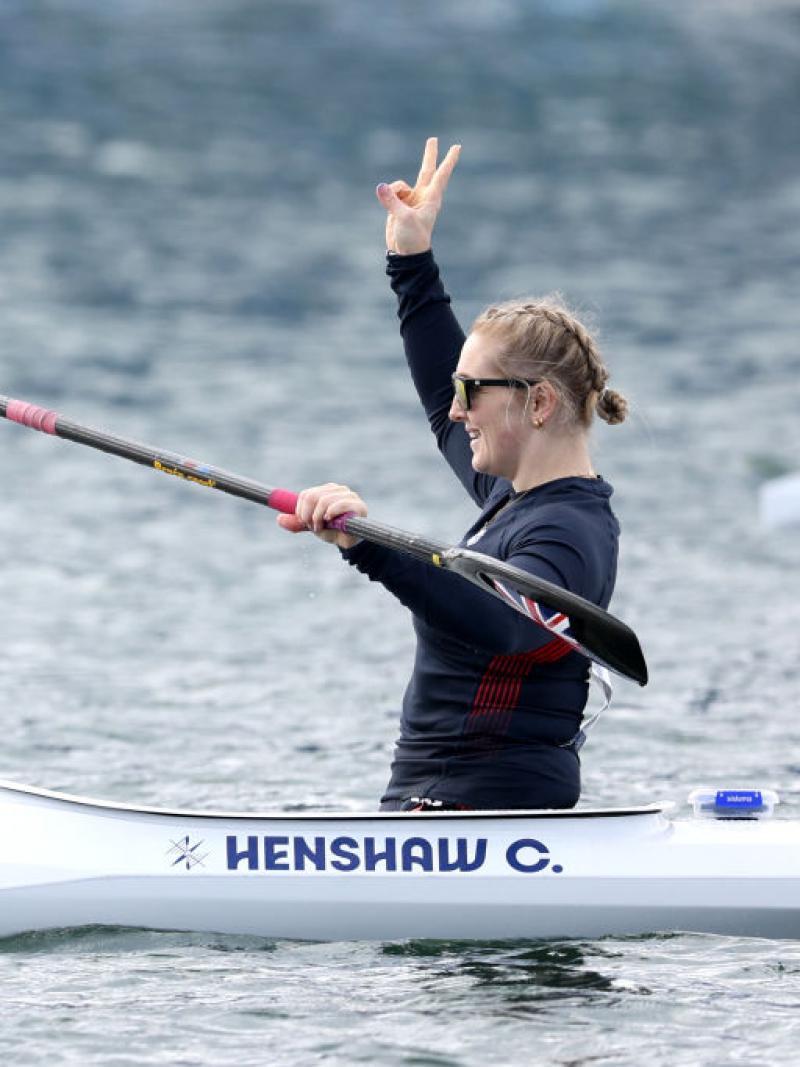A woman in a canoe holds her oats with one hand and lifts the other hand in the air, making a peace sign