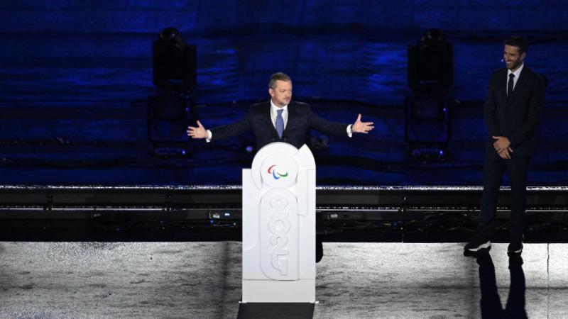 A man on a stage raises his arms and stands behind a podium that is decorated with the year 2024 and the Paralympic agitos