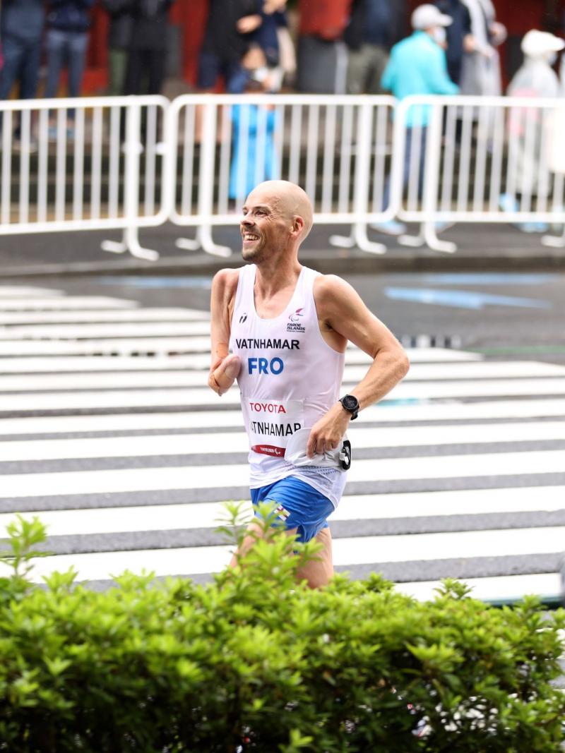 An athlete who is missing his right arm below the elbow smiles as he runs on the street during a marathon as onlookers take photos of him.