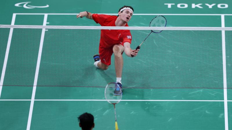 A Para badminton match between two male athletes at Tokyo 2020. A player in red uniform prepares to return the shuttle near the net.
