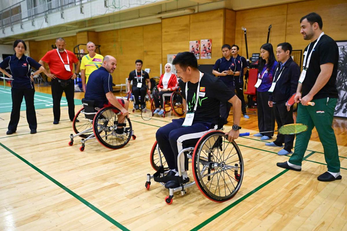 Two men holding badminton racquets are demonstrating on wheelchairs in front of 10 people