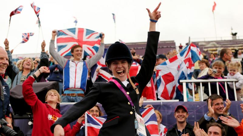 A female Para equestrian athlete poses for a photograph in front of the stands. Spectators are holding the GB flag